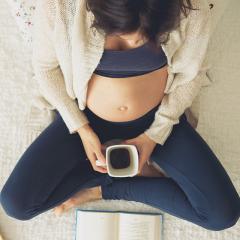 A pregnant woman sits comfortably on the floor with a black coffee and book